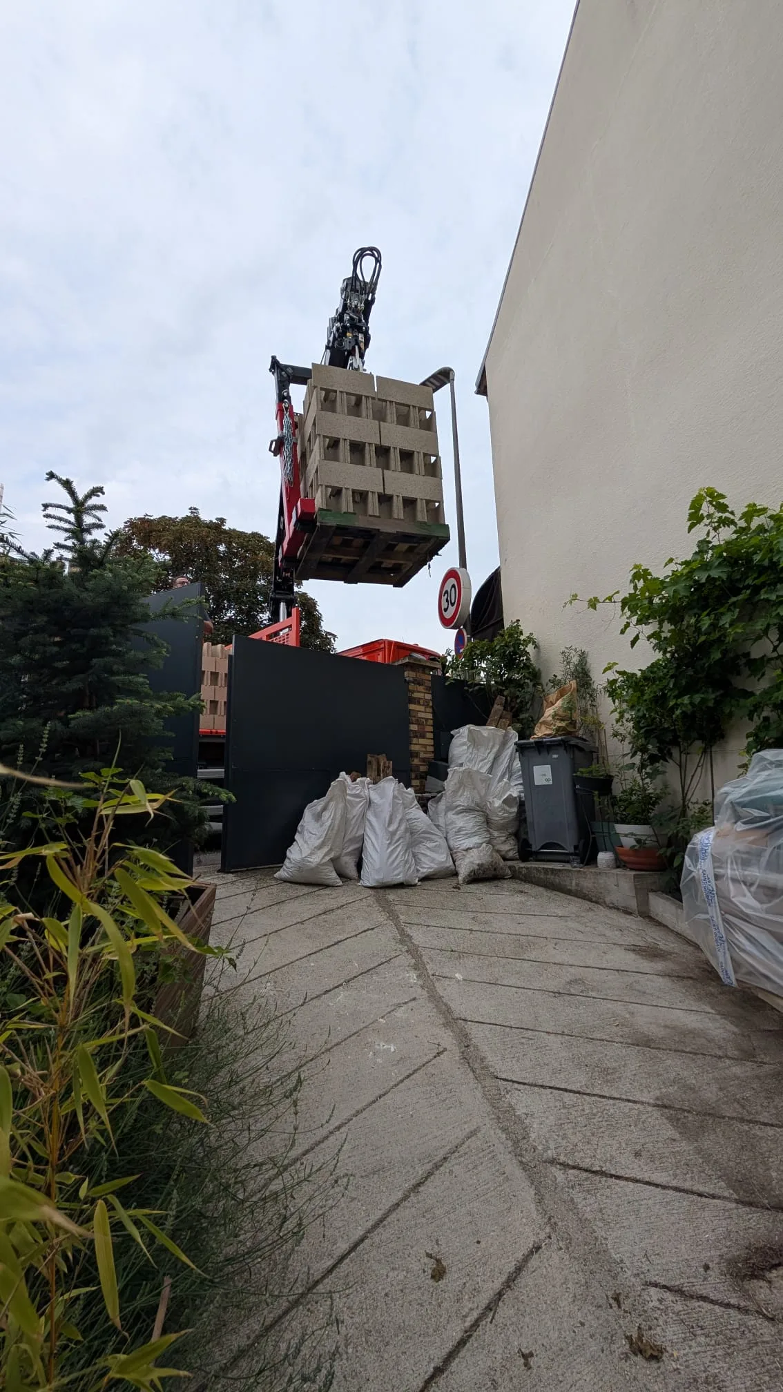 Palette de parpaings livrée à la grue par-dessus le mur, big bags de mélange posés au sol à l'entrée de la cour