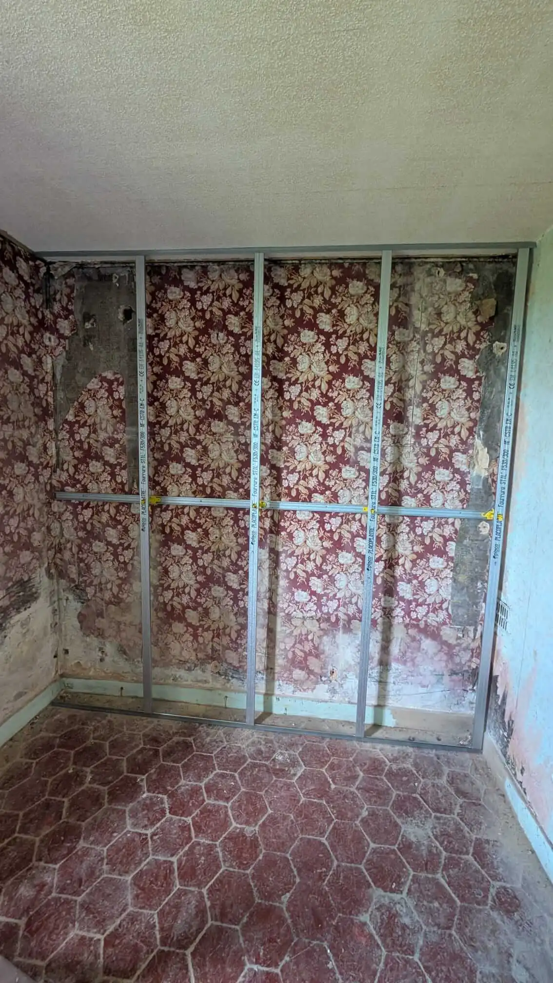 Ossature Optima posée sur un mur d'une chambre, avec le papier peint fleuri d'origine encore en place